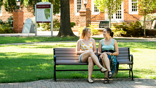 Two students sit on a bench and chat on the Quad.