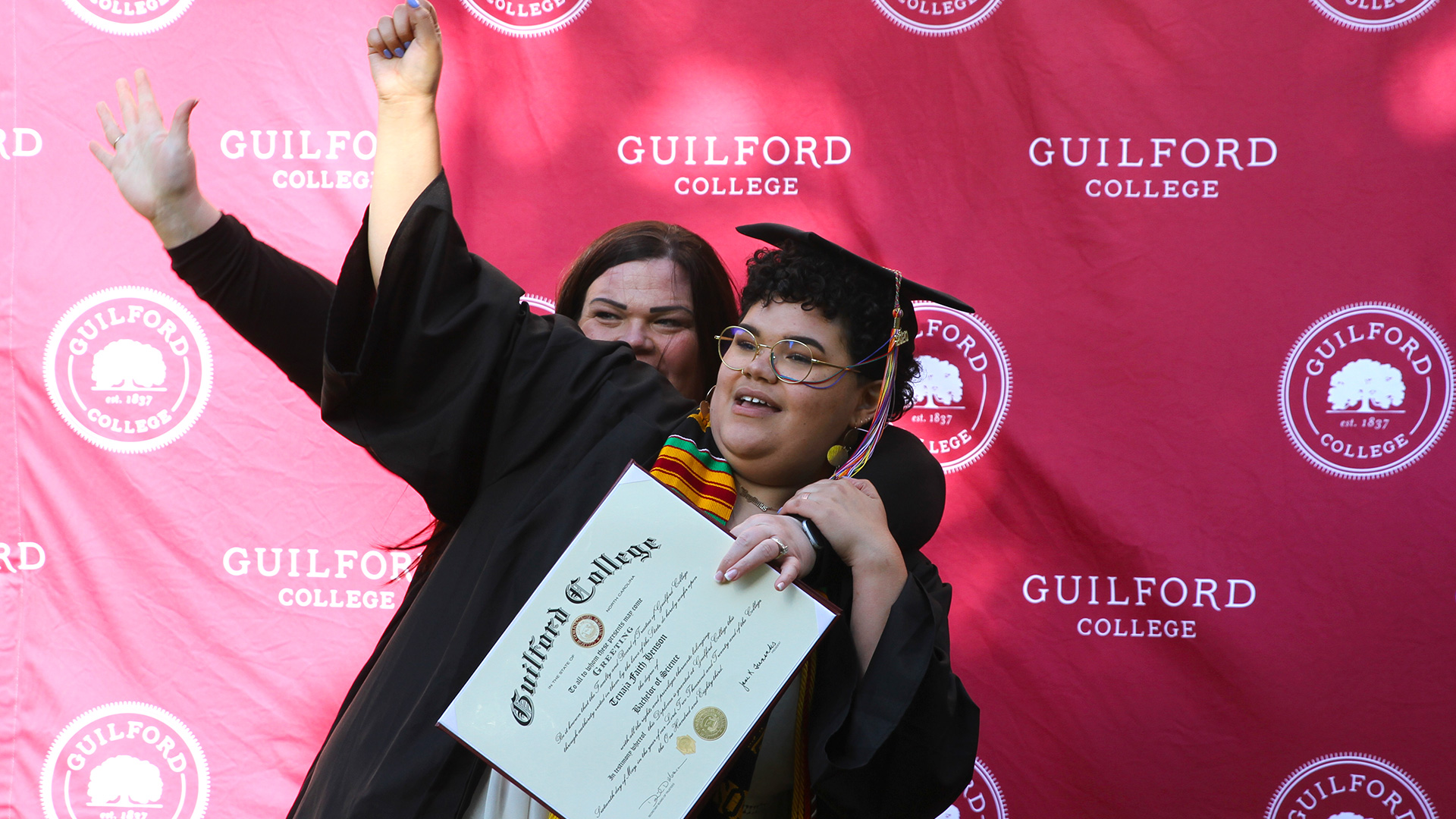 A graduate celebrates with a friend in front of the Guilford College step-and-repeat.