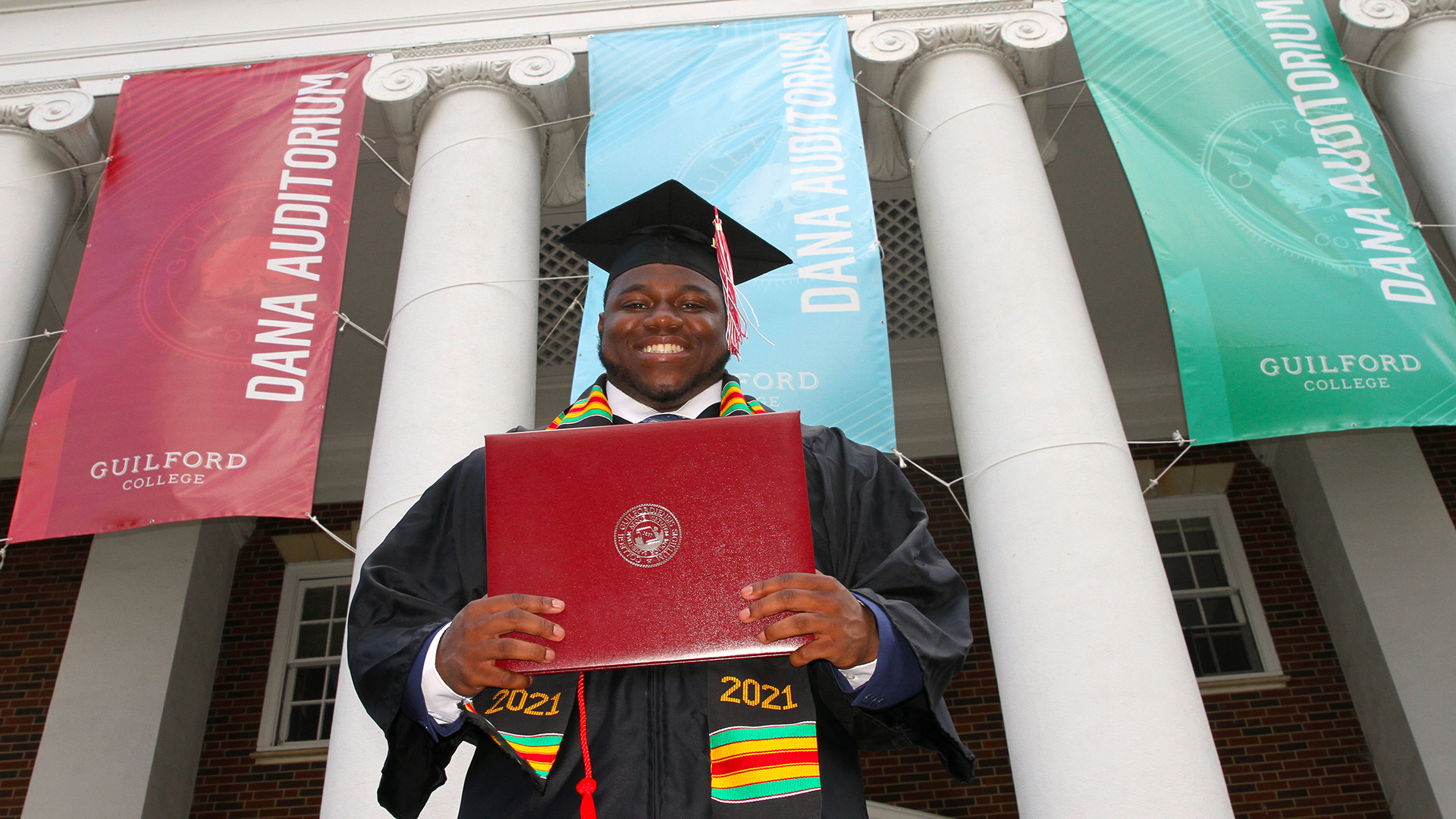 A proud 2021 graduates holds his diploma in front of Dana Auditorium.