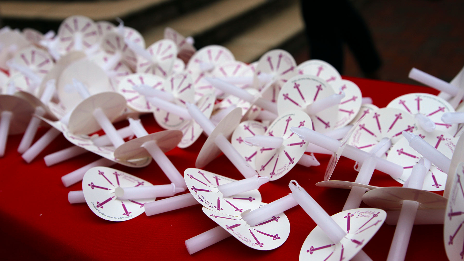 A pile of white candles is on a table covered in a red tablecloth.