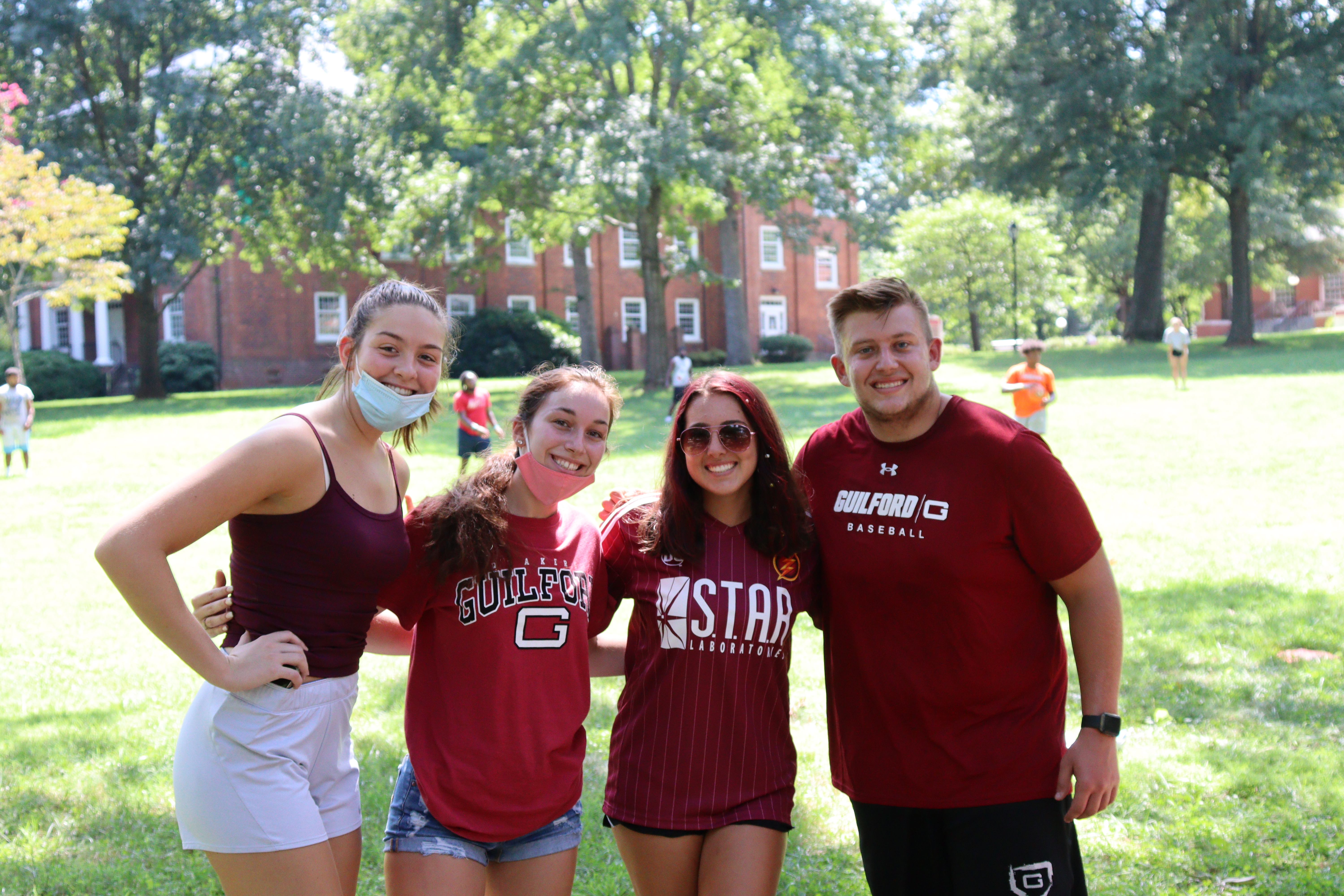 Four students wearing Guilford t-shirts (and one with a burgundy tank top) stand together for a group photo on the Quad.