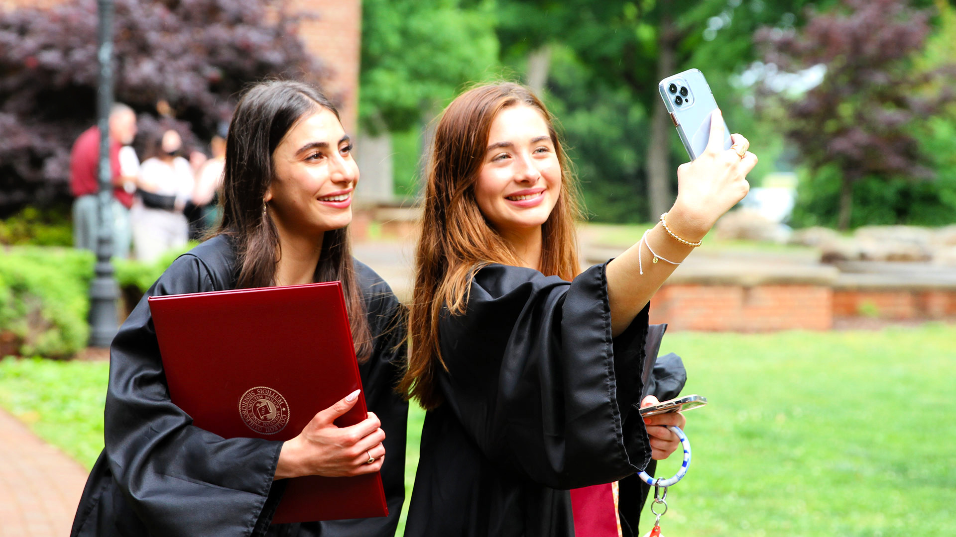 A student holds up a phone to take a photo with another graduate outdoors after Commencement.