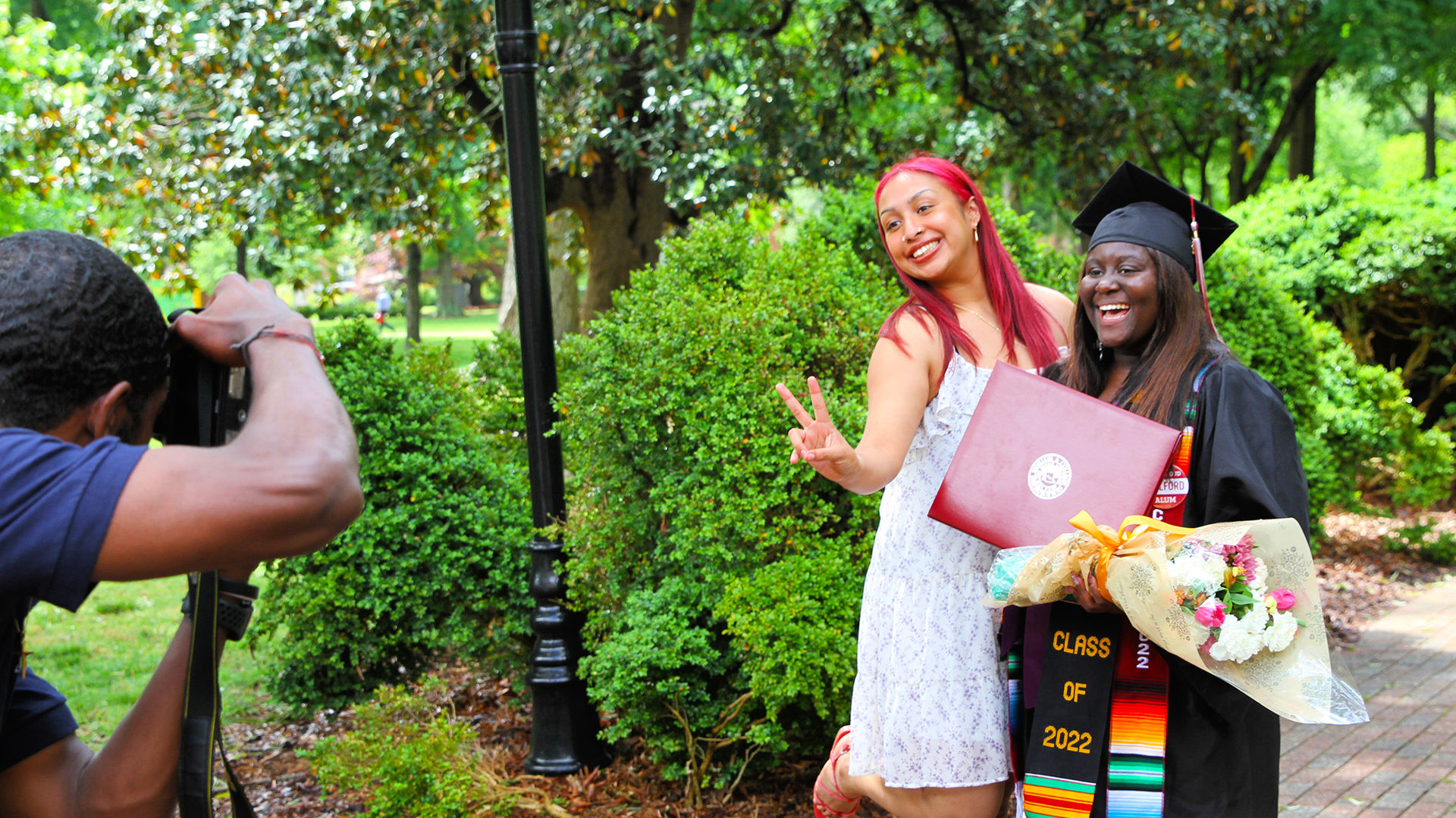 A friend takes a photo with a graduate wearing a graduation cap and gown outdoors after Commencement.