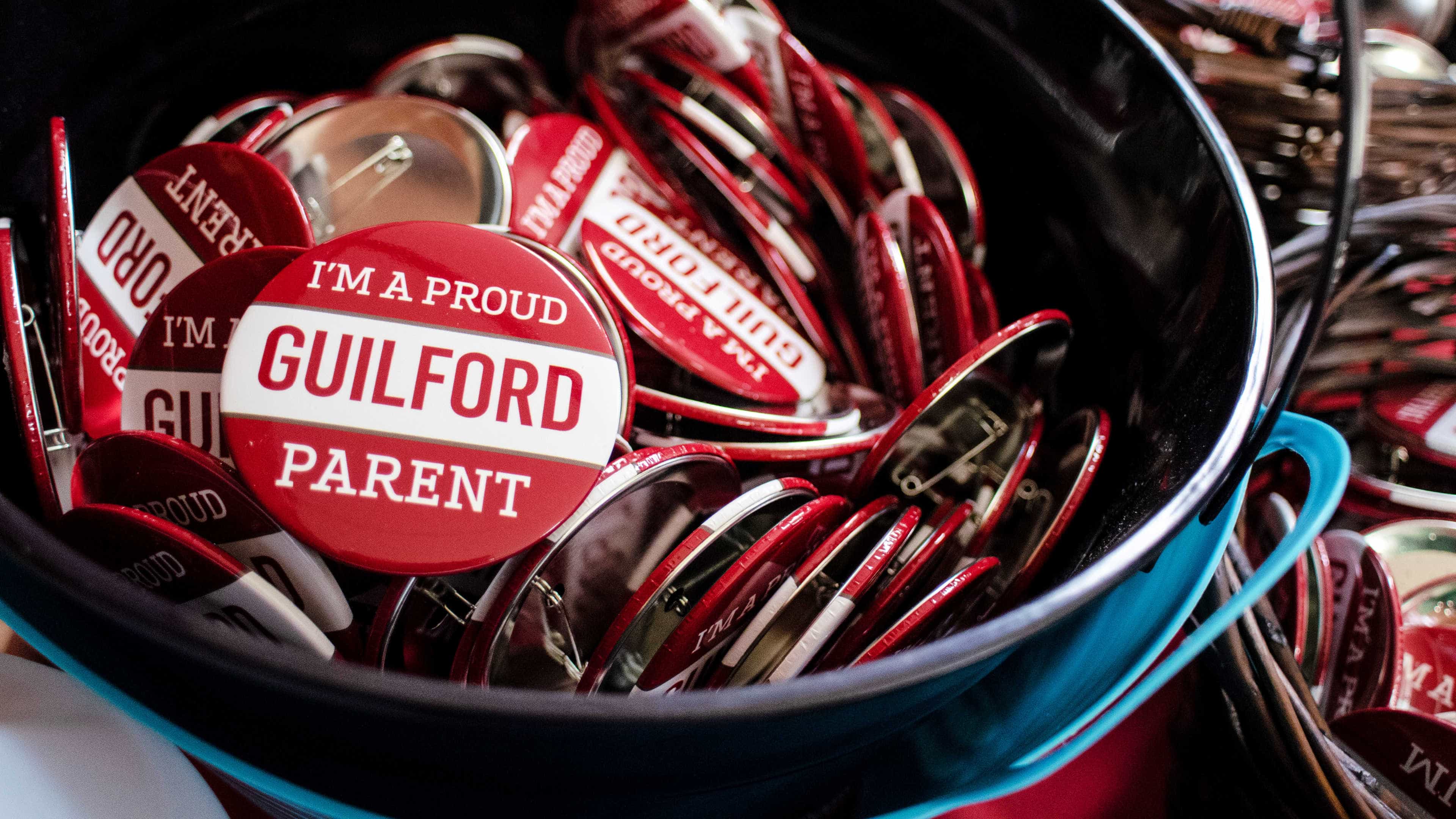 A bucket is filled with red and white pins that read, I'm a Proud Guilford Parent.