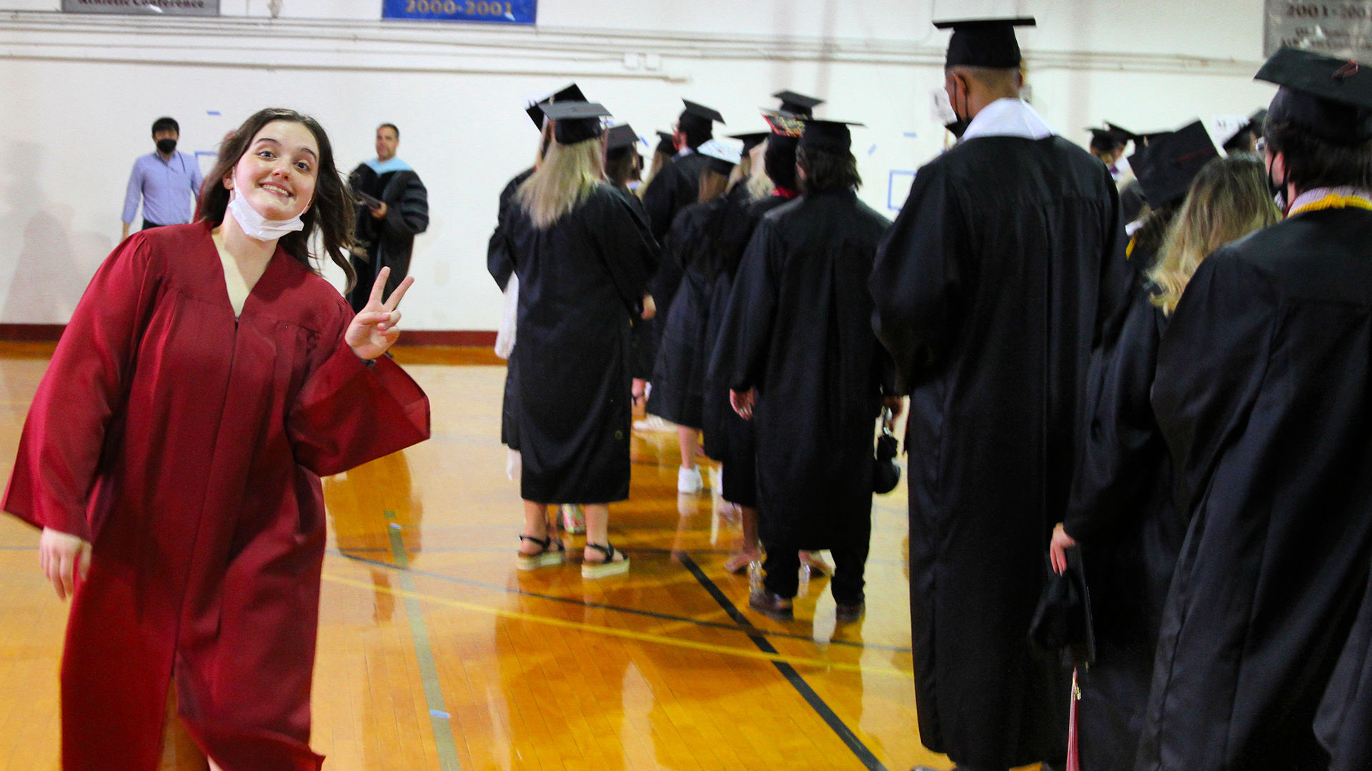 A junior marshal wearing a red robe gives a peace sign to the camera as they walk by lined-up graduates.