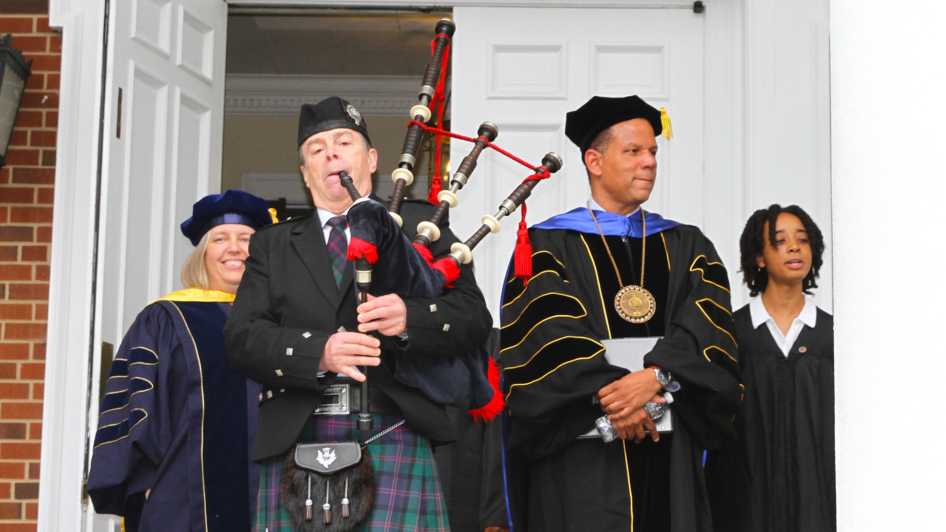A bagpiper leads Provost Maria Rosales, President Kyle Farmbry, and Student Body President Tinyah Erin '23 onto Dana's porch, while other attendees file out behind them.