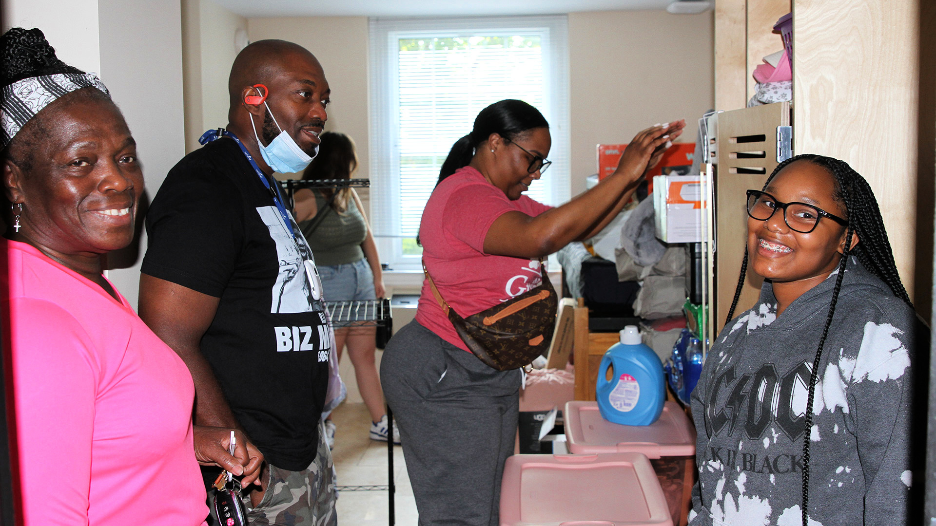 A student and friend smile at the camera, as other friends and family members place items in the student's res-hall room.