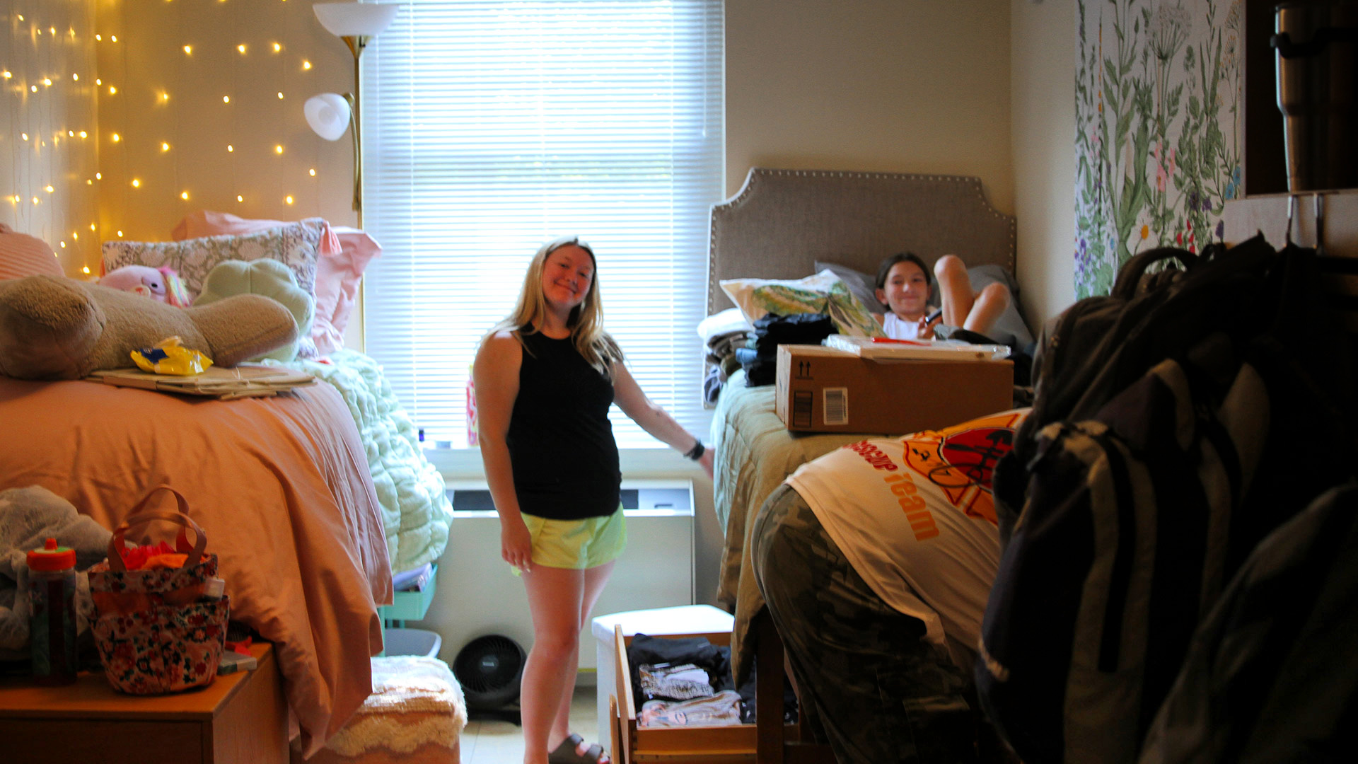 A student stands between two beds in a residence hall and smiles at the camera, while their roommates smiles from their bed.