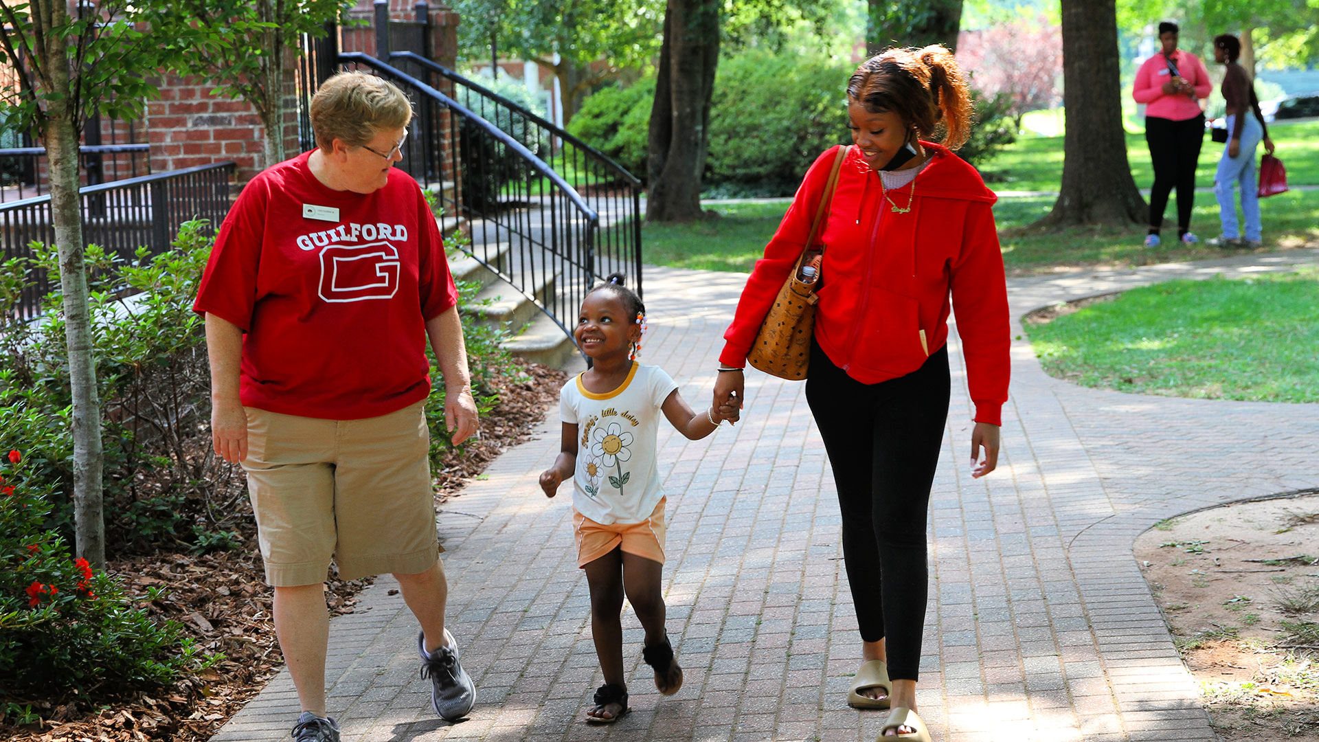 A staff member wearing a Guilford t-shirt walks and chats with a visitor and a small child on the Quad.