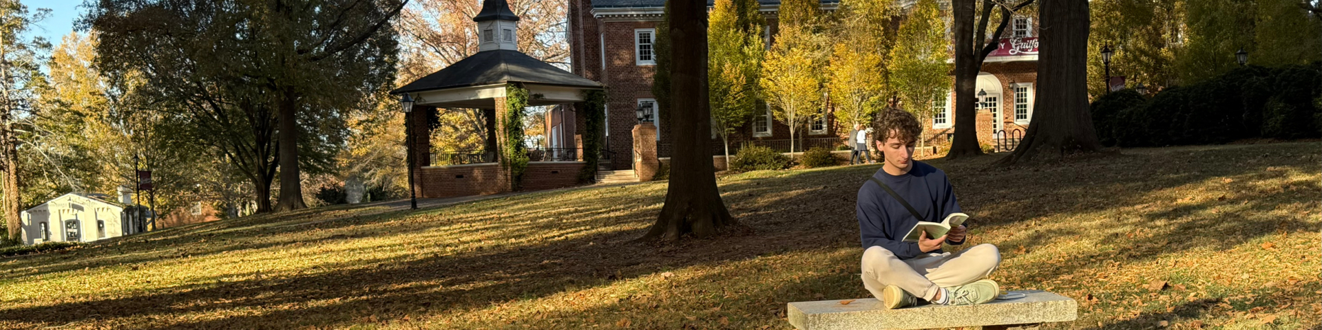 Guilford College student reads a book on a campus bench on a sunny fall day.