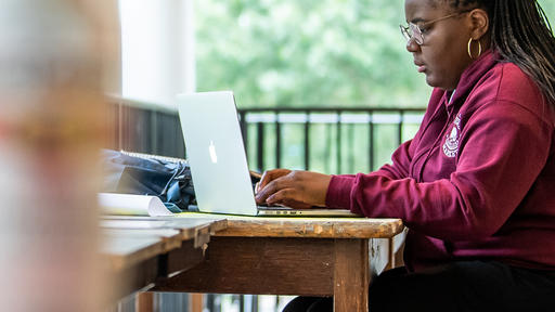 A Guilford student wearing a long sleeve red sweatshirt works outside on a MacBook computer.