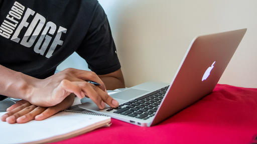 A student wearing a black shirt that says Guilford Edge works on a MacBook computer.