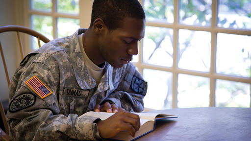 A student wearing military fatigues reads a book at a table in the library.