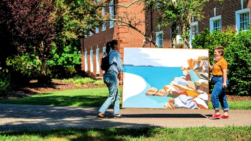 Two students carry a large painting showing an ocean and a rock cliff.