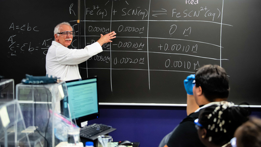 A Biology professor wearing safety goggles stands at a blackboard and speaks to seated students.