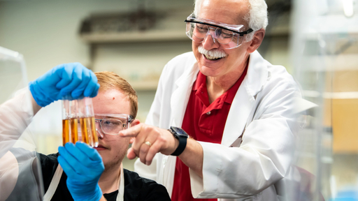 A Biology professor wearing goggles helps a student review four test tubes.