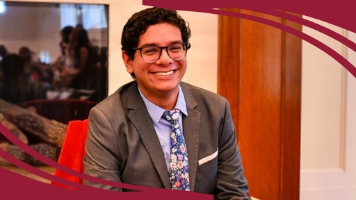 Jose Jacobo, wearing a gray blazer, blue shirt, and tie, sits indoors on a red chair.