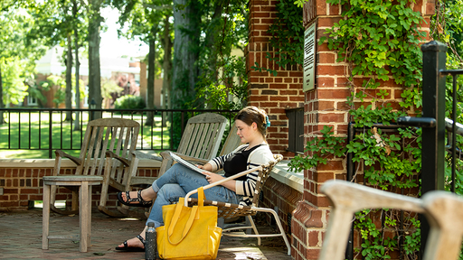 A student sits in a chair on Founders Hall Patio, reading a book, with a large yellow bag beside their chair.