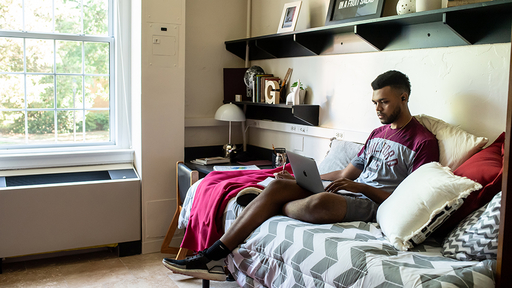 A student sits on a residence-hall bed and reads.