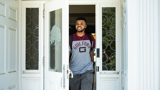 A student wearing a Guilford shirt and backpack smiles while walking out of the door to a residence hall.
