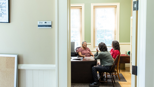 Two students chat with a professor in their office on campus.