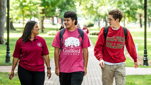 A staff member walks down the Quad brick walkway with two students wearing red Guilford shirts.
