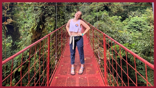 Drew Erickson stands on a red suspension bridge, surrounded by trees, wearing long blue pants, a cropped tank top, a shirt around the waist, and sandals.