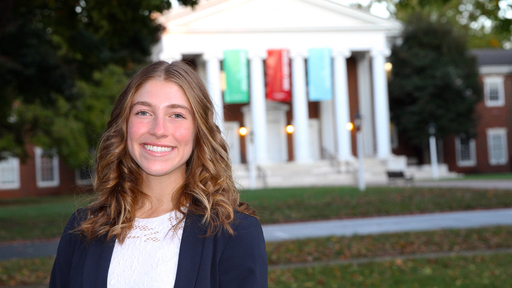 Grace Calus stands outdoors in front of Dana Auditorium, wearing a white shirt under a blue jacket.