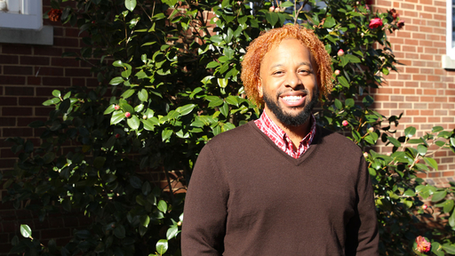 Quinton standing in front of King hall, wearing a brown v-neck sweater