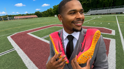 Smiling Guilford athlete in a suit holds track spikes on the campus athletic field.