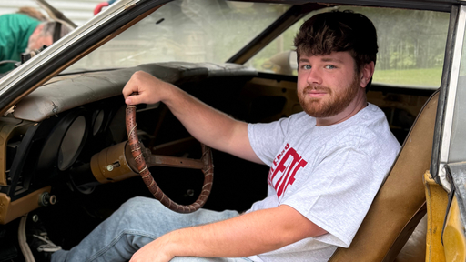 Guilford College student in gray tee smiles from driver's seat of a vintage car.