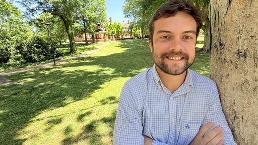 Smiling man leaning on a tree on the sunny Guilford College campus.