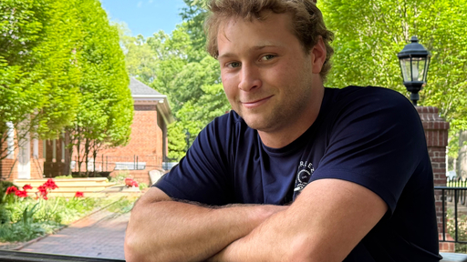 Smiling young man in a blue shirt with crossed arms on Guilford College campus.