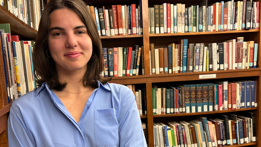 Smiling student in a blue shirt, surrounded by books in a Guilford College library.