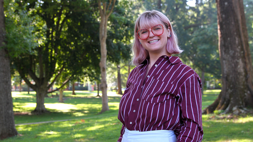Rachel is posing in the academic quad, wearing a burgundy stripped blouse and glasses.. 