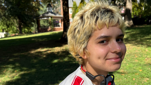 Young person with short, blonde hair smiling gently, with a gazebo on Guilford campus in background.
